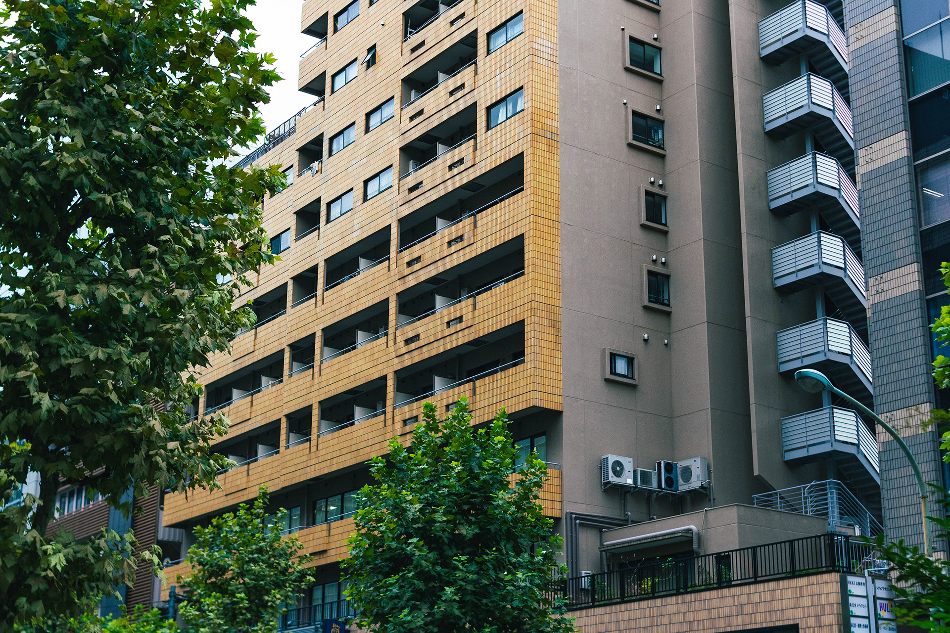 Photograph of a residential concrete building with a facade decorated with slightly reflective yellow tiles and a trees lining the left and bottom of the foreground.